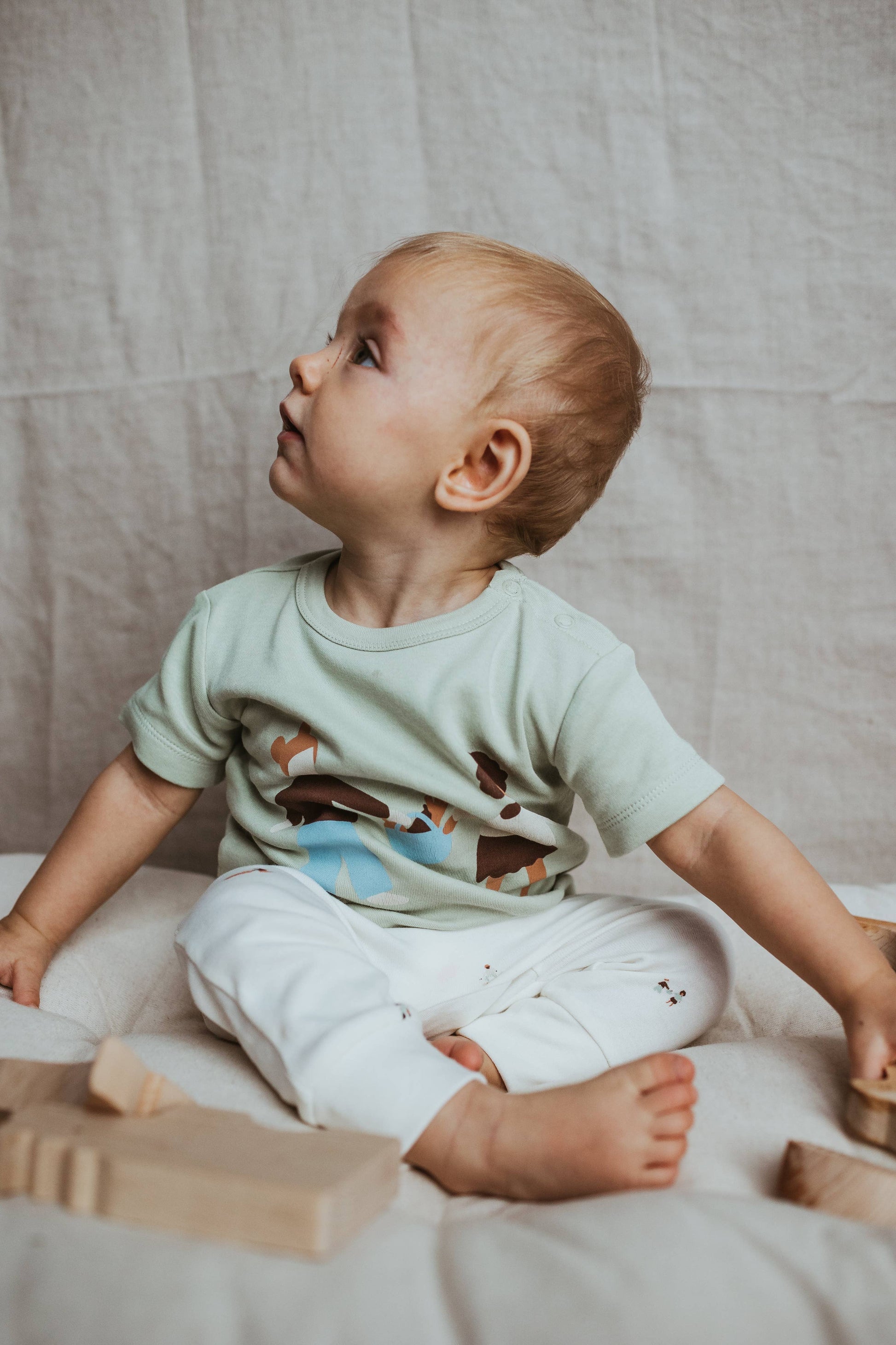 Baby sitting on a bed with wooden blocks, wearing a light green shirt and white pants.