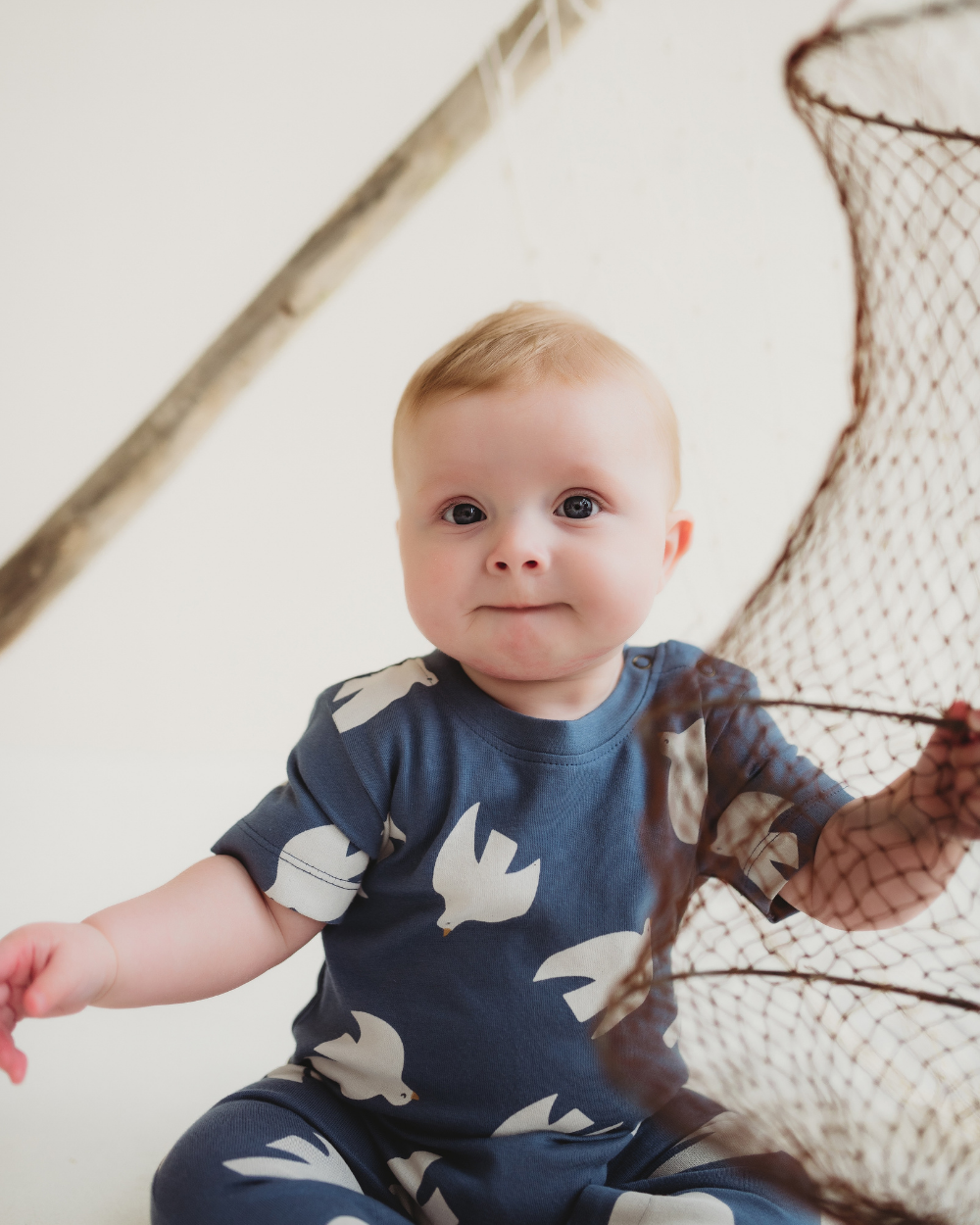Baby wearing a blue onesie with bird patterns, sitting next to a fishing net.