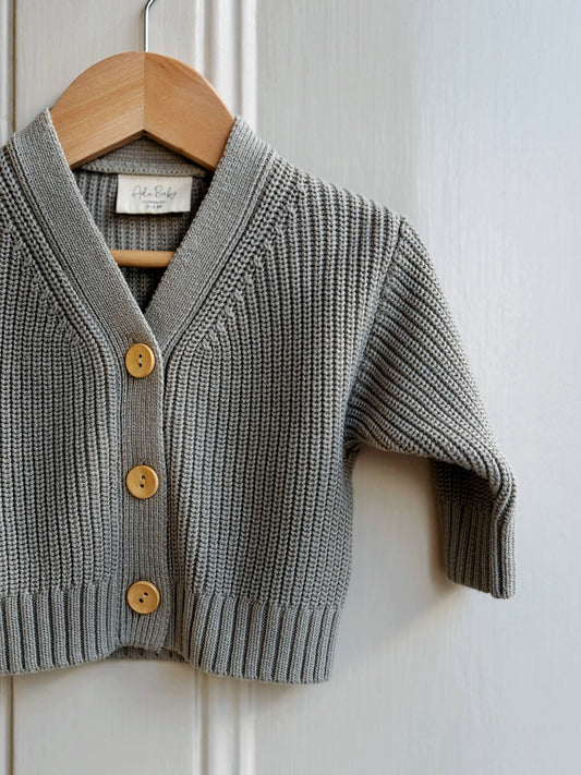 Gray knitted cardigan with gold buttons on a wooden hanger against a white paneled wall.