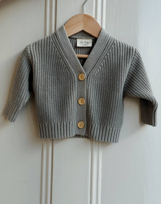Gray knitted cardigan with gold buttons on a wooden hanger against a white paneled wall.