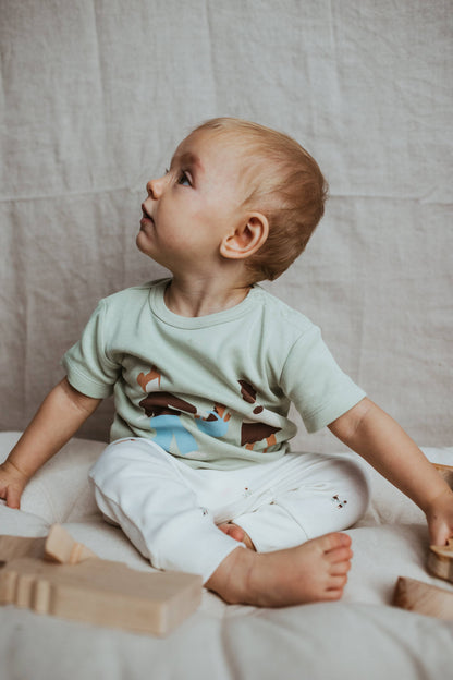 Baby sitting on a bed with wooden blocks, wearing a light green shirt and white pants.