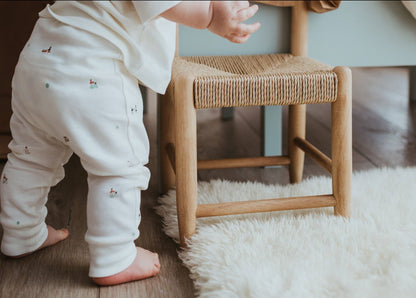 Child in white pants standing next to a wooden stool on a fluffy rug