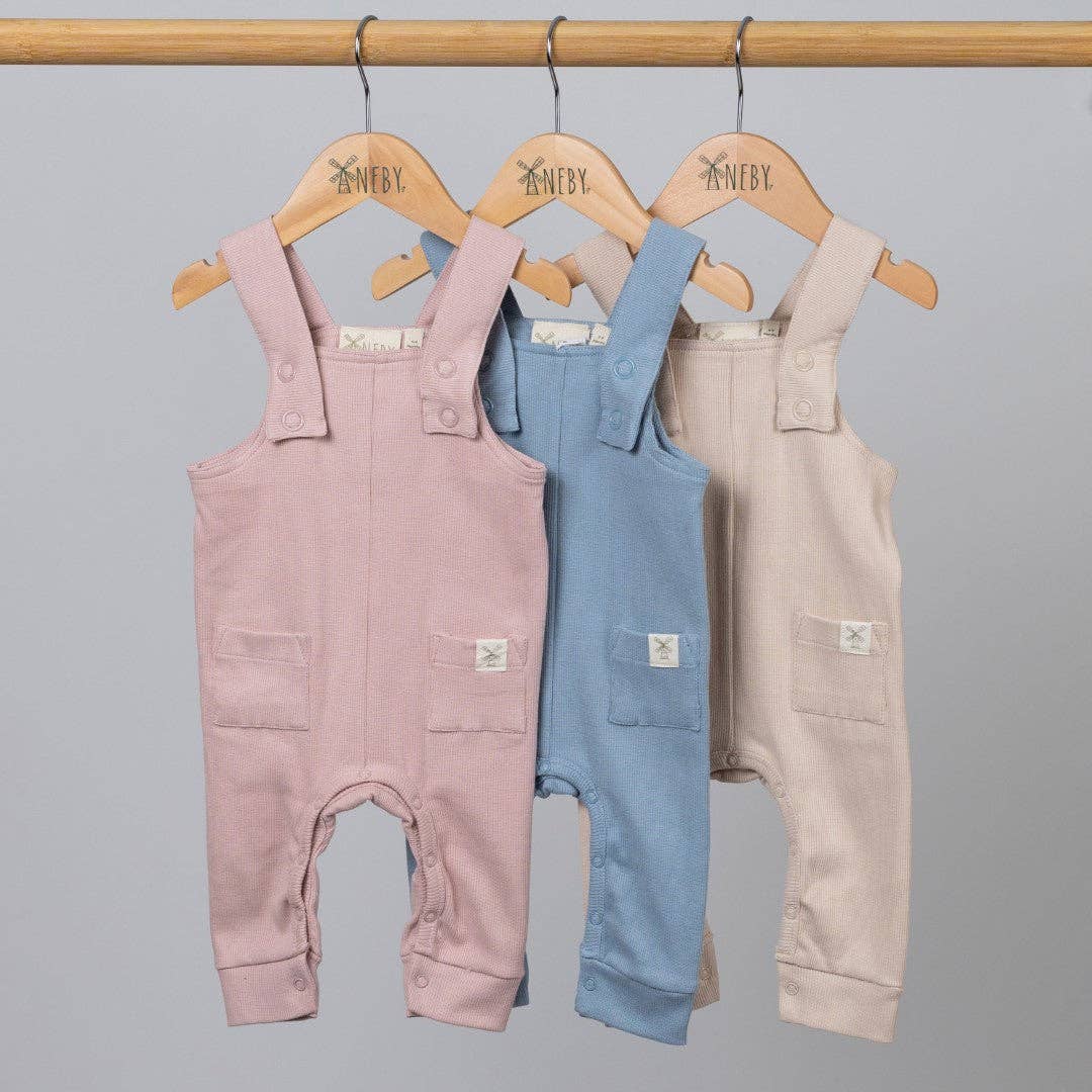 Three children's dungarees in pink, blue, and beige hanging on a rack against a gray background.
