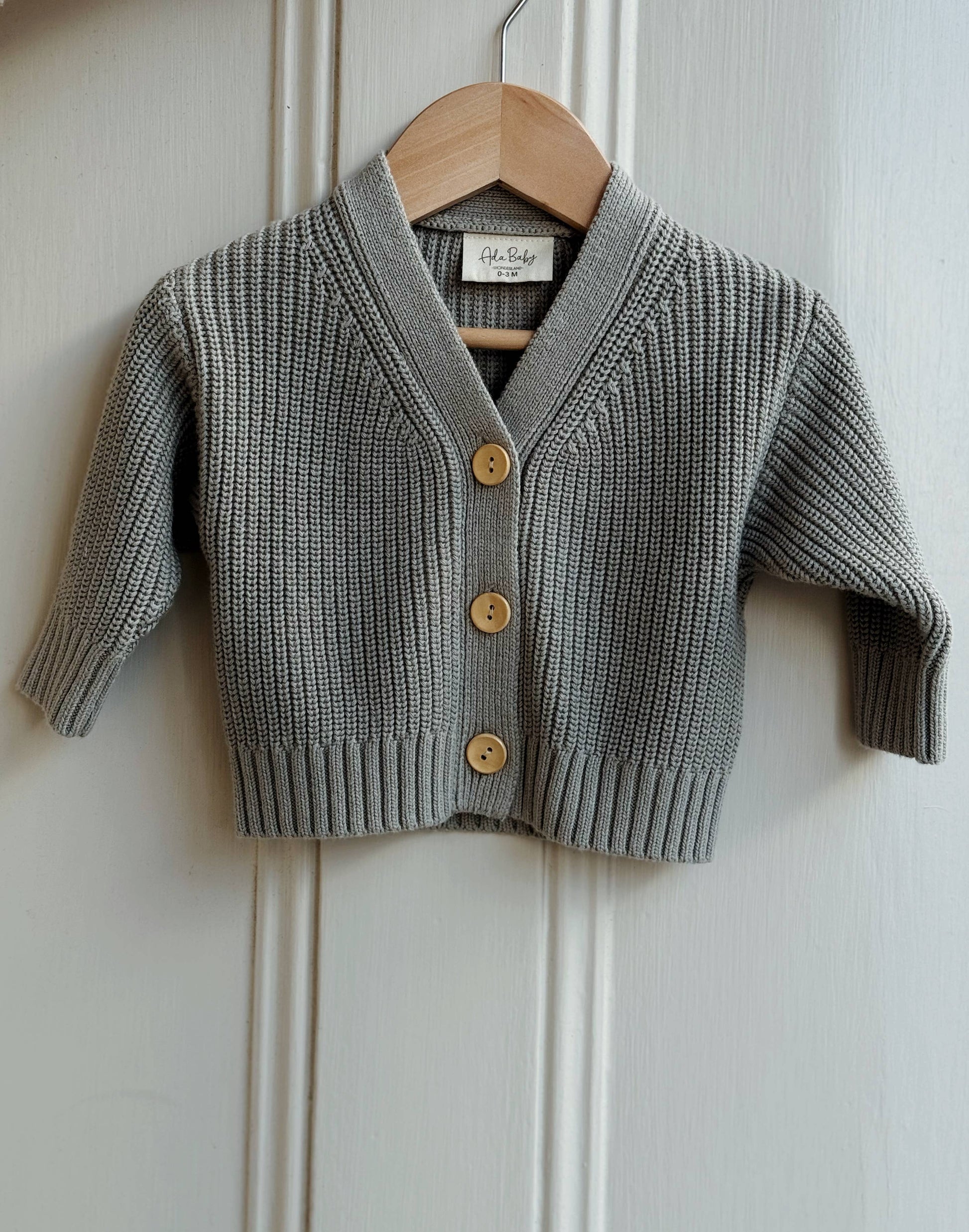 Gray knitted cardigan with gold buttons on a wooden hanger against a white paneled wall.