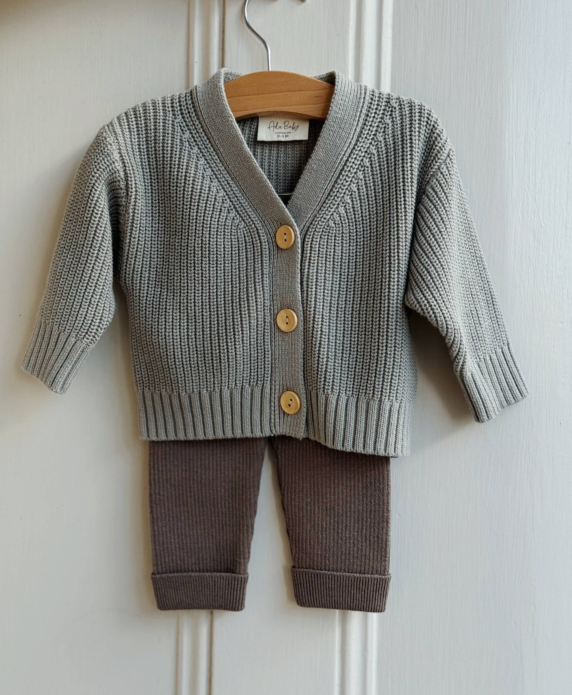 Children's gray knitted cardigan and brown pants set on a hanger against a white wall.