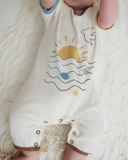 Baby wearing a white romper with sun and wave design, lying on a textured surface.