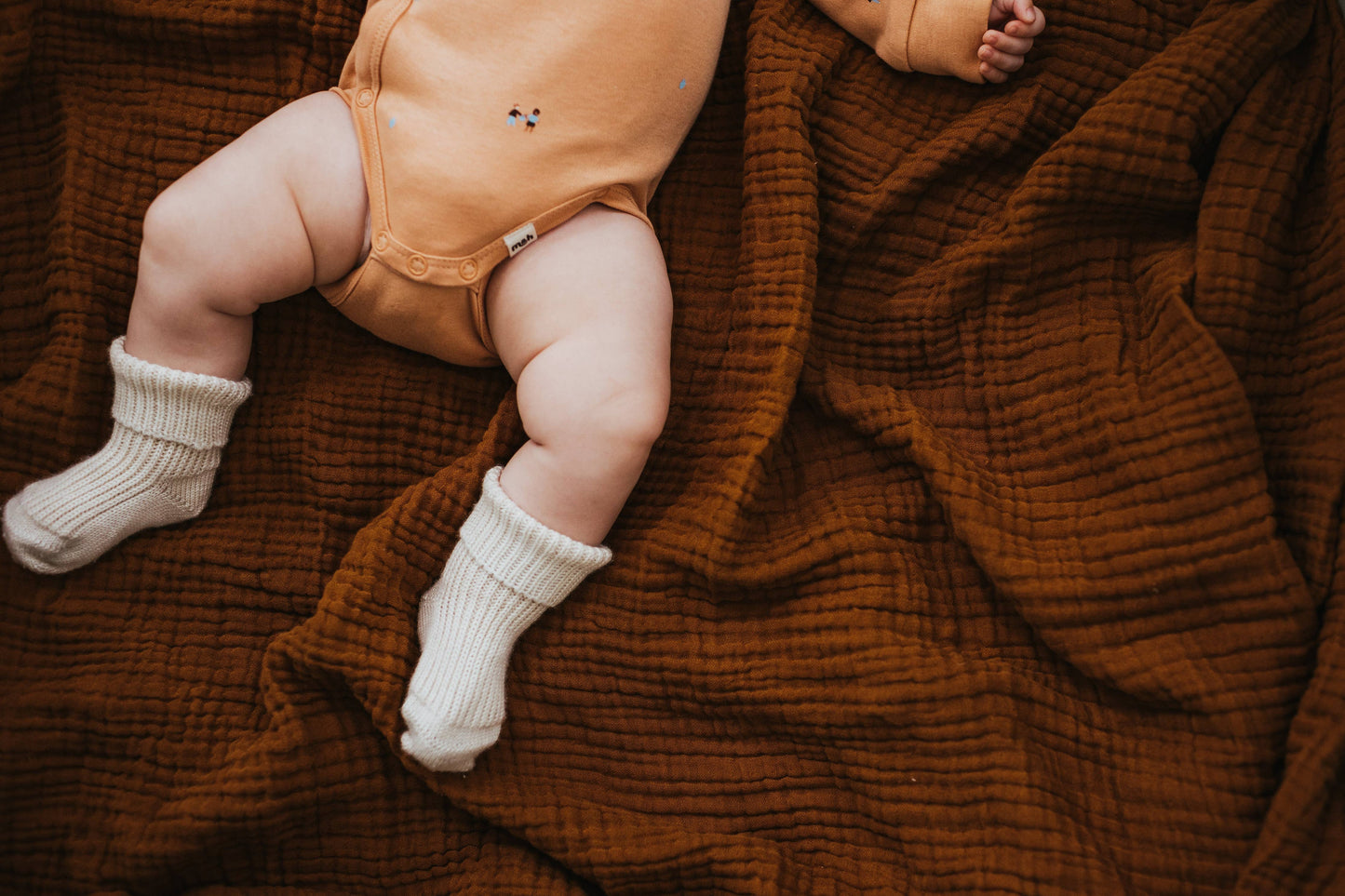 Baby in a tan outfit and white socks sitting on a brown textured blanket
