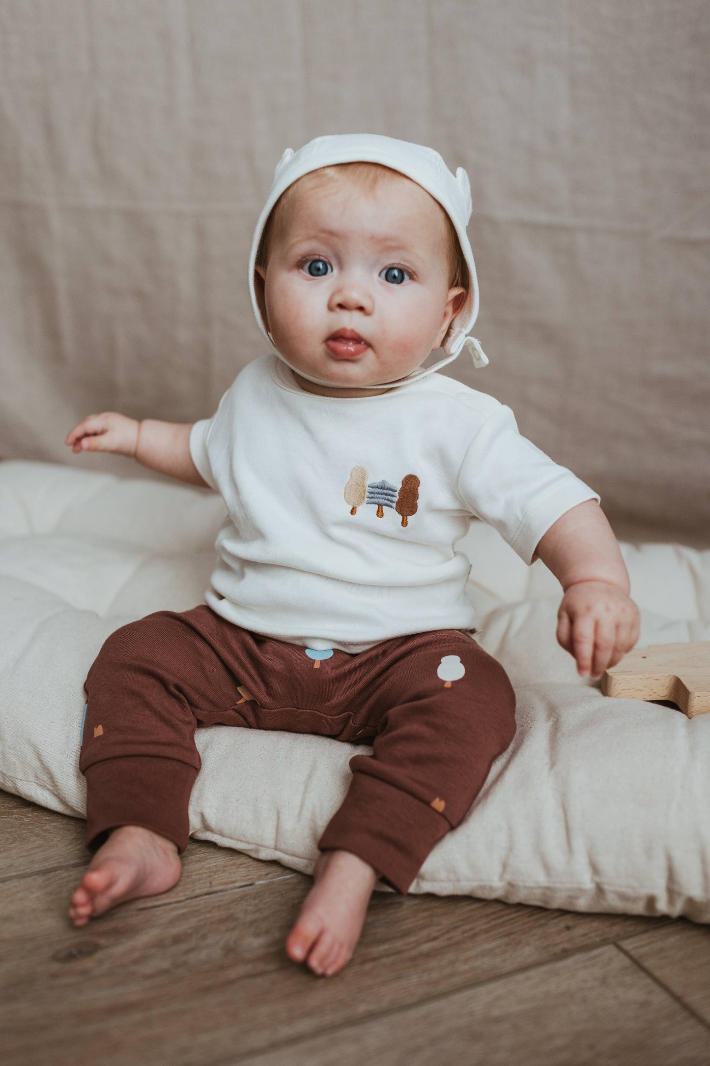 Baby wearing a white shirt with cartoon characters and brown pants, sitting on a beige cushion.