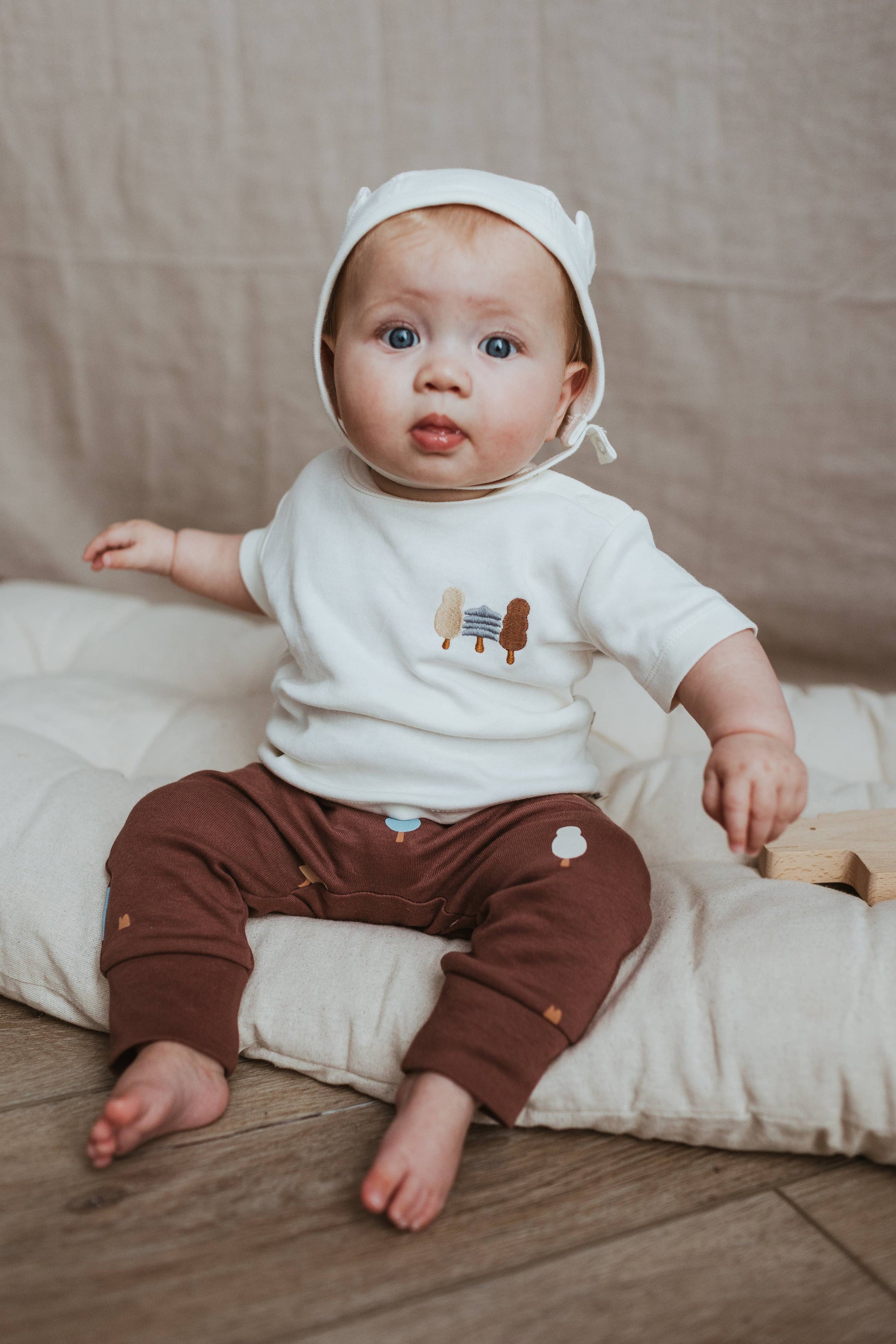 Baby wearing a white shirt with cartoon characters and brown pants, sitting on a beige cushion.