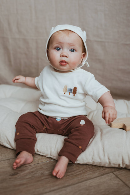 Baby wearing a white shirt with cartoon characters and brown pants, sitting on a beige cushion.